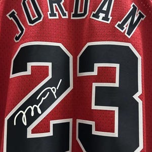 Peut inclure: Un maillot de basket rouge avec le nom JORDAN en lettres noires &agrave; bordure blanche au-dessus du num&eacute;ro 23 en noir &agrave; bordure blanche. Une signature blanche est inscrite sur le num&eacute;ro 23 &agrave; l'avant du v&ecirc;tement en tissu maill&eacute;.