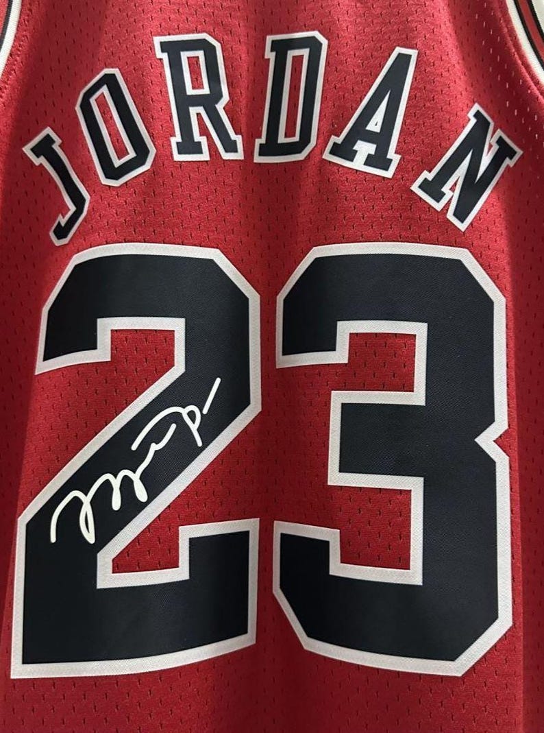 Peut inclure: Un maillot de basket rouge avec le nom JORDAN en lettres noires &agrave; bordure blanche au-dessus du num&eacute;ro 23 en noir &agrave; bordure blanche. Une signature blanche est inscrite sur le num&eacute;ro 23 &agrave; l'avant du v&ecirc;tement en tissu maill&eacute;.