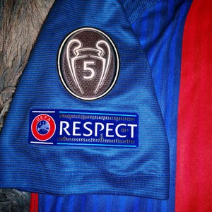 May include: A blue soccer jersey with a red and yellow stripe on the right side. The jersey has a patch with a UEFA Champions League trophy and the number 5 on it. The jersey also has a patch that says "UEFA RESPECT".