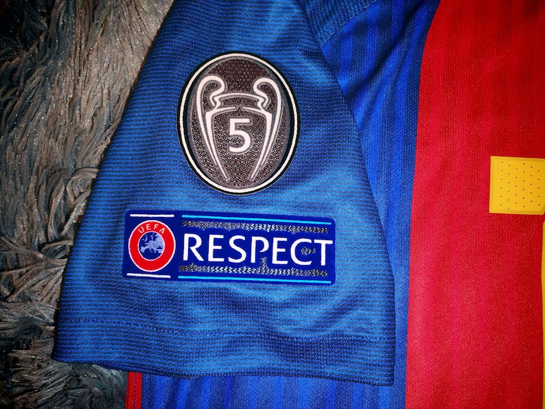 May include: A blue soccer jersey with a red and yellow stripe on the right side. The jersey has a patch with a UEFA Champions League trophy and the number 5 on it. The jersey also has a patch that says "UEFA RESPECT".