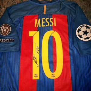May include: A blue soccer jersey with red and yellow stripes. The number 10 is printed on the back with the name "MESSI" above it. The jersey has a Champions League patch on the right sleeve and a patch that says "RESPECT" on the left sleeve.