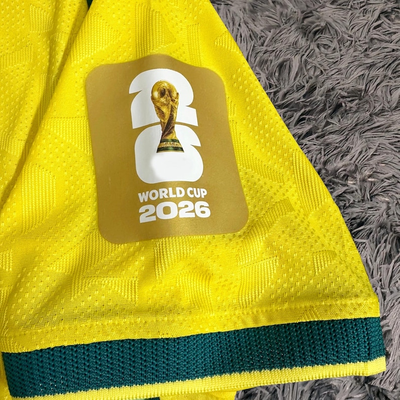 May include: A bright yellow athletic jersey sleeve featuring a textured geometric pattern and a dark green hem. A rectangular patch displays the white text 26, the FIFA World Cup trophy graphic, and the words WORLD CUP 2026 in white lettering.