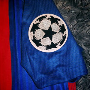 May include: A blue soccer jersey with a red and yellow stripe on the side. The jersey has a Champions League patch on the sleeve.