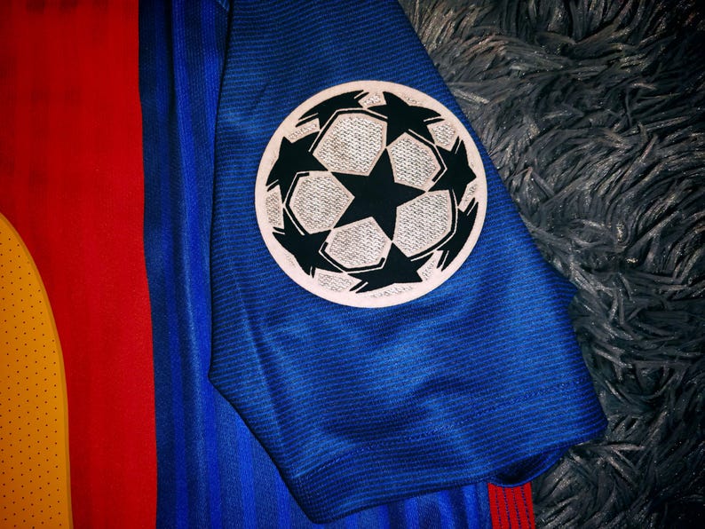 May include: A blue soccer jersey with a red and yellow stripe on the side. The jersey has a Champions League patch on the sleeve.