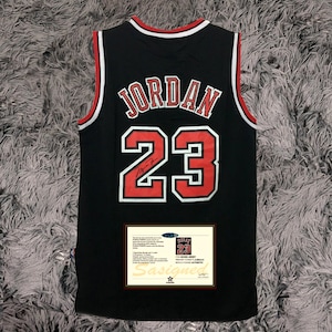 以下が含まれることがあります： 黒色のバスケットボールジャージ。名前「JORDAN」と番号「23」が赤と白で表示されています。ジャージは赤と白のトリムが付いています。真正性の証明書が添付されています。