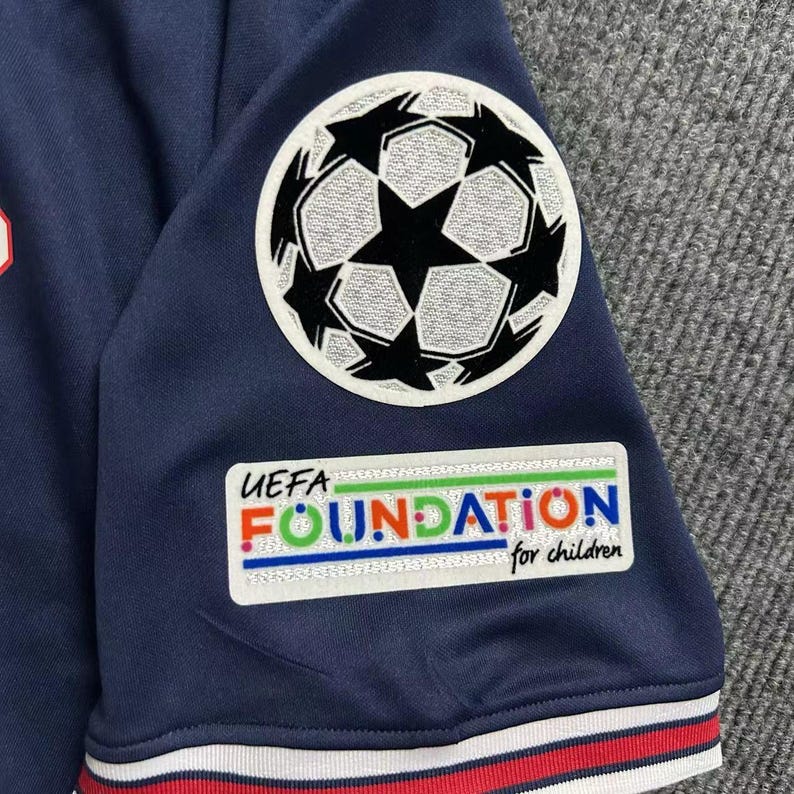May include: Navy blue soccer jersey with a white, black, and red trim. The jersey features a UEFA Foundation patch and a soccer ball logo. The text "UEFA FOUNDATION for children" is visible on the jersey.