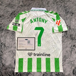 May include: A white and green soccer jersey with the name "ANTONY" and the number "7" printed on the back. The jersey has green stripes and logos, including "REVEL" and "trainline". A framed certificate is also visible.
