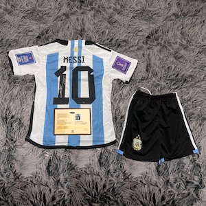May include: Argentina soccer jersey and shorts set. The jersey is white with light blue stripes, the number 10, and the name MESSI on the back. The shorts are black with white stripes and a team logo. A certificate is also included.