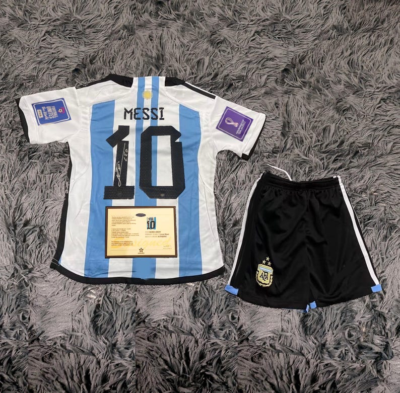 May include: Argentina soccer jersey and shorts set. The jersey is white with light blue stripes, the number 10, and the name MESSI on the back. The shorts are black with white stripes and a team logo. A certificate is also included.