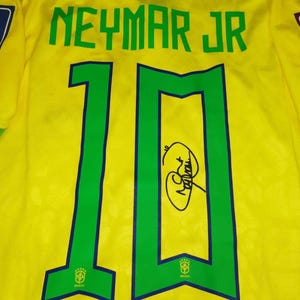 May include: A yellow soccer jersey featuring the name NEYMAR JR and the number 10 in green with blue outlines. The jersey includes a FIFA World Cup Qatar 2022 patch, a Football Unites the World patch, and a black ink signature on the number 10.