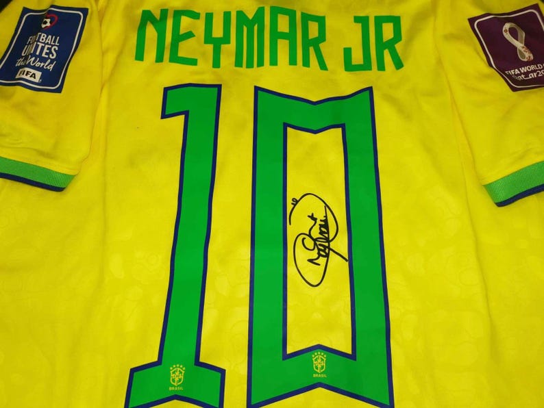 May include: A yellow soccer jersey featuring the name NEYMAR JR and the number 10 in green with blue outlines. The jersey includes a FIFA World Cup Qatar 2022 patch, a Football Unites the World patch, and a black ink signature on the number 10.