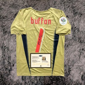Pode incluir: Uma camisa de futebol dourada com detalhes pretos e a palavra "buffon" impressa nas costas. A camisa tem uma faixa vermelha e branca nas costas com uma assinatura em preto. A camisa tem um pequeno patch na manga direita com o logotipo da Copa do Mundo da FIFA.