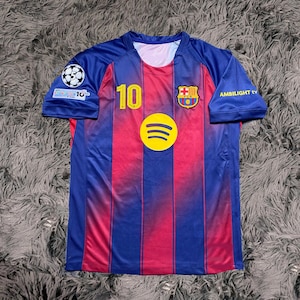 May include: A blue and red soccer jersey with the number 10 in yellow. The jersey features the Spotify logo, a team crest, and the text "AMBILIGHT tv". The jersey has a round neck and short sleeves.