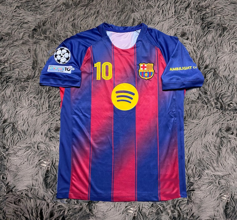 May include: A blue and red soccer jersey with the number 10 in yellow. The jersey features the Spotify logo, a team crest, and the text "AMBILIGHT tv". The jersey has a round neck and short sleeves.