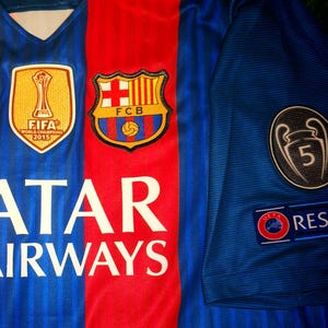 May include: A blue and red soccer jersey with the team logo, "FCB", and the text "Qatar Airways". The jersey also features a FIFA World Champions 2015 patch and a UEFA Champions League patch with the number 5.