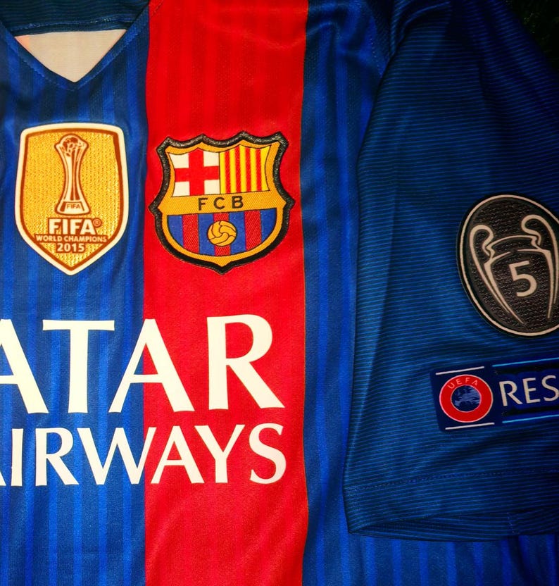 May include: A blue and red soccer jersey with the team logo, "FCB", and the text "Qatar Airways". The jersey also features a FIFA World Champions 2015 patch and a UEFA Champions League patch with the number 5.