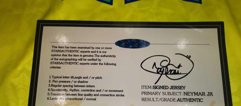 May include: A rectangular certificate of authenticity with a black border on a bright yellow background. The document features the text STARSAUTHENTIC, a holographic seal, a signature, and the text ITEM: SIGNED JERSEY, PRIMARY SUBJECT: NEYMAR JR, RESULT/GRADE: AUTHENTIC.