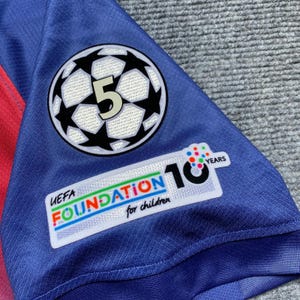 May include: Close-up of a blue soccer jersey with a white and black soccer ball logo featuring the number 5. The jersey also has a patch that says "UEFA FOUNDATION for children 10 YEARS".