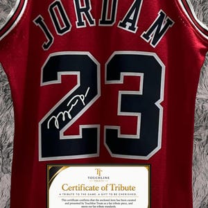 Peut inclure: Un maillot de basket rouge portant le nom JORDAN et le num&eacute;ro 23 en noir avec des contours blancs. Une signature blanche est visible sur le chiffre 2. Un certificat de tribut rectangulaire de Touchline Treats est plac&eacute; en bas.