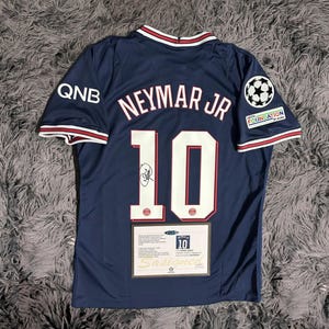Pode incluir: Camisola de futebol azul marinho com o nome "Neymar Jr" e o número "10" em branco. A camisola tem uma assinatura e patches. Tem acabamentos vermelhos e brancos na gola e nas mangas. O logótipo QNB está na manga esquerda.