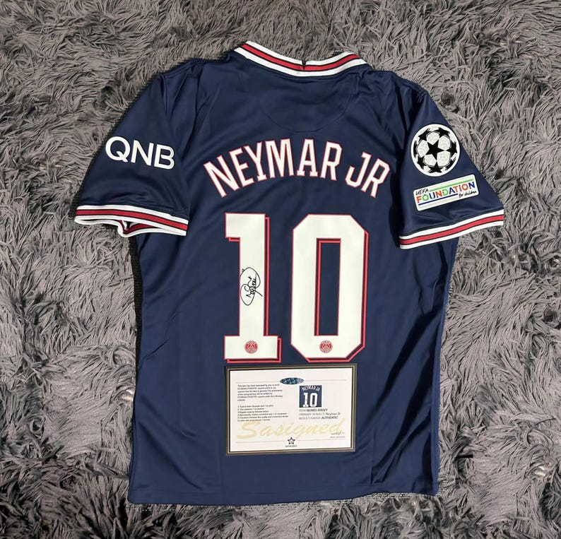 May include: Navy blue soccer jersey with the name "Neymar Jr" and the number "10" in white. The jersey has a signature and patches. The jersey has red and white trim on the collar and sleeves. The jersey has the QNB logo on the left sleeve.