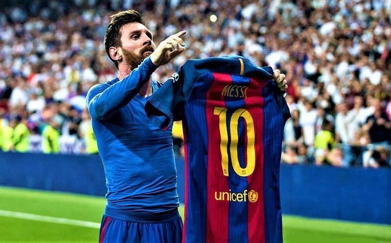 May include: A soccer player in a blue jersey holds up a red and blue soccer jersey with the number 10 and the name "MESSI" on the back. The jersey also has the UNICEF logo and the text "unicef".