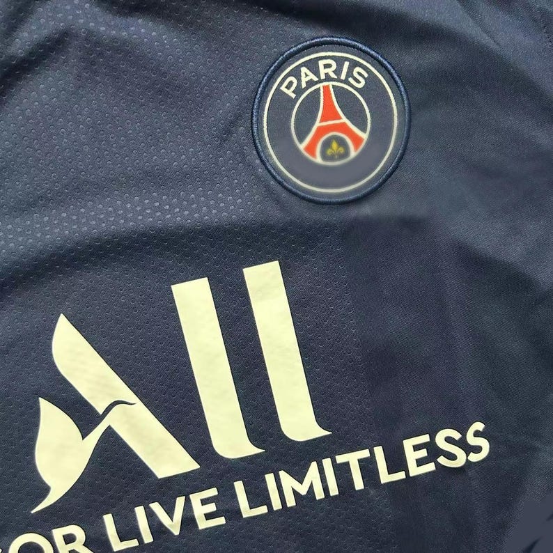 May include: Close-up of a navy blue jersey with the Paris Saint-Germain logo patch. The logo is a circular emblem with the Eiffel Tower and the word "PARIS". The word "ALL" and the phrase "OR LIVE LIMITLESS" are printed in white.