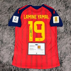 May include: A red soccer jersey with dark blue sleeves, featuring the name LAMINE YAMAL and the number 19 in yellow. The jersey includes a black signature, a Unite For Peace patch, a World Cup 2026 patch, and an authentication certificate label.