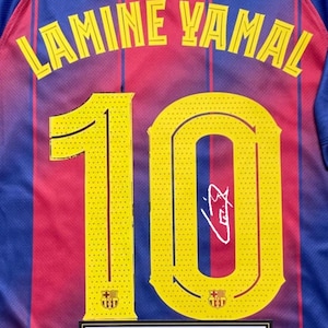 May include: A close-up of a blue, red, and yellow soccer jersey. The jersey features the name "LAMINE YAMAL" in yellow lettering across the back, with the number "10" in yellow. A signature is visible on the number. The FC Barcelona crest is present.