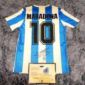 Puede incluir: Una camiseta de fútbol azul y blanca a rayas con el nombre "MARADONA" y el número "10" en la espalda. La camiseta está firmada con tinta negra.