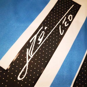 May include: Close-up of a jersey with a blue and white striped design. A black band with white polka dots and a white signature is visible. The jersey has a light blue background.
