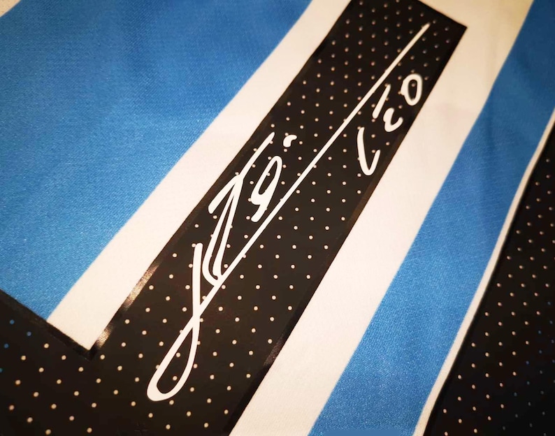 May include: Close-up of a jersey with a blue and white striped design. A black band with white polka dots and a white signature is visible. The jersey has a light blue background.