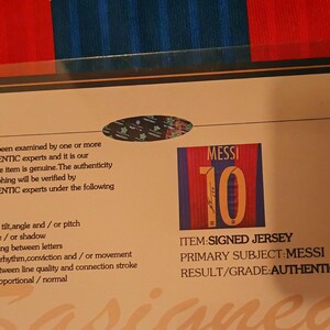 May include: A white card with a blue and red striped background. The card has a picture of a soccer jersey with the number 10 and the name "MESSI" on it. The card also has text that says "ITEM SIGNED JERSEY", "PRIMARY SUBJECT MESSI", and "RESULT/GRADE AUTHENTIC".