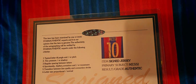 May include: A white card with a blue and red striped background. The card has a picture of a soccer jersey with the number 10 and the name "MESSI" on it. The card also has text that says "ITEM SIGNED JERSEY", "PRIMARY SUBJECT MESSI", and "RESULT/GRADE AUTHENTIC".