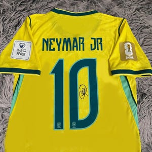 May include: A yellow Brazil soccer jersey with green trim, featuring the name NEYMAR JR and the number 10 in dark green. The back includes a signature on the number, a Unite For Peace patch on the right sleeve, and a World Cup 2026 patch on the left.
