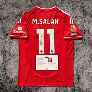 May include: Red soccer jersey with white accents, featuring the name "M. SALAH" and the number "11" on the back. Includes sponsor logos and a "No room for racism" patch. A signed certificate is attached.