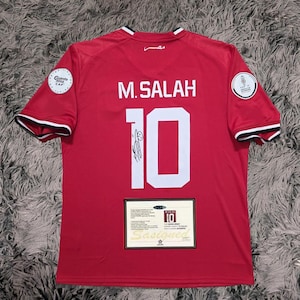 May include: Red football shirt with "M. SALAH" and the number "10" on the back. The shirt has white and black trim on the sleeves and collar. Includes a certificate of authenticity and patches.