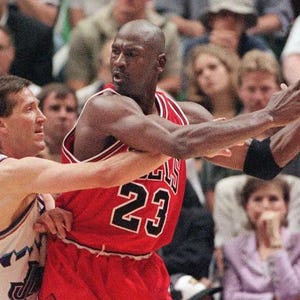 Peut inclure: Un joueur de basket portant un maillot rouge avec le num&eacute;ro 23 en noir et blanc, tenant un ballon de basket en cuir orange. Un second joueur en maillot blanc avec des bordures bleues et vertes est positionn&eacute; tout pr&egrave;s, face &agrave; la m&ecirc;me direction.