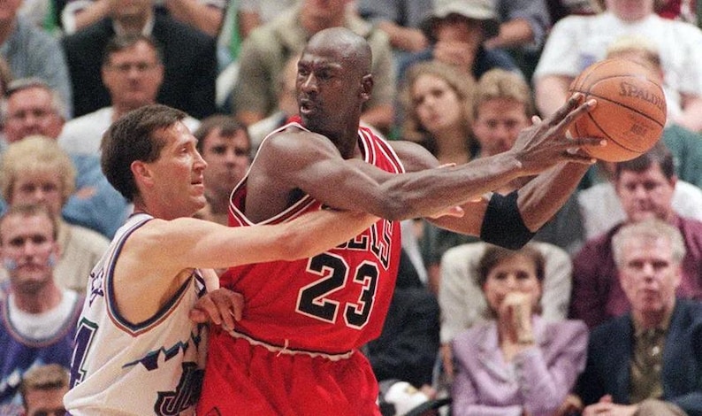 Peut inclure: Un joueur de basket portant un maillot rouge avec le num&eacute;ro 23 en noir et blanc, tenant un ballon de basket en cuir orange. Un second joueur en maillot blanc avec des bordures bleues et vertes est positionn&eacute; tout pr&egrave;s, face &agrave; la m&ecirc;me direction.