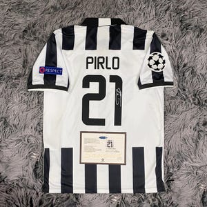 Puede incluir: Una camiseta de fútbol a rayas blancas y negras con el nombre "PIRLO" y el número "21" impresos en la espalda. La camiseta tiene un parche "RESPECT" y un logotipo de la Liga de Campeones. Se muestra un certificado de autenticidad debajo.