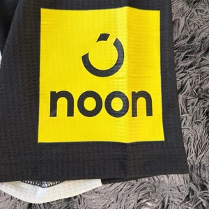 Może przedstawiać: Zbliżenie czarnej koszulki sportowej z ż&oacute;łtym kwadratowym logo. Logo przedstawia zakrzywiony wz&oacute;r nad słowem "noon" w czarnej, pogrubionej czcionce. Koszulka ma wzorzystą fakturę i białe akcenty.