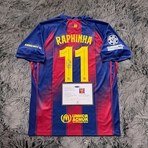May include: A blue and red soccer jersey with the name "RAPHINHA" and the number "11" in yellow. The jersey has a signature and a certificate of authenticity. The UNHCR ACNUR logo is visible at the bottom.