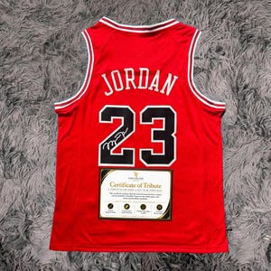 Peut inclure: Un maillot de basket rouge avec le nom JORDAN et le num&eacute;ro 23 en noir avec des contours blancs. Une signature noire est visible sur le num&eacute;ro 23. Une &eacute;tiquette rectangulaire Certificate of Tribute est fix&eacute;e sur la partie inf&eacute;rieure avant.