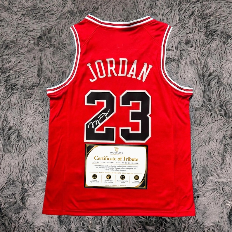 Peut inclure: Un maillot de basket rouge avec le nom JORDAN et le num&eacute;ro 23 en noir avec des contours blancs. Une signature noire est visible sur le num&eacute;ro 23. Une &eacute;tiquette rectangulaire Certificate of Tribute est fix&eacute;e sur la partie inf&eacute;rieure avant.