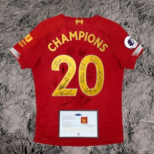Op de afbeelding: Rood voetbalshirt met gouden letters "CHAMPIONS 20". Het shirt heeft meerdere handtekeningen en een Premier League-badge. Een certificaat van echtheid is zichtbaar.
