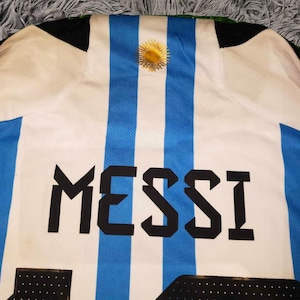 May include: Argentina soccer jersey with blue and white stripes, featuring the name "MESSI" in bold black letters. A gold emblem is centered at the top. The jersey's design is a classic representation of the Argentinian national team.