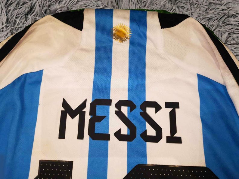 May include: Argentina soccer jersey with blue and white stripes, featuring the name "MESSI" in bold black letters. A gold emblem is centered at the top. The jersey's design is a classic representation of the Argentinian national team.