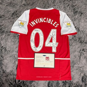 Op de afbeelding: Een rood-wit voetbalshirt met het woord "INVINCIBLES" op de rug. Het nummer "04" is zichtbaar, samen met handtekeningen van spelers. De mouwen zijn wit met een Champions 2003-2004 logo. Een certificaat van echtheid is inbegrepen.