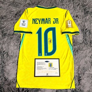 May include: A yellow Brazil soccer jersey featuring the name NEYMAR JR and the number 10 in dark green with a black signature on the number. The jersey includes a Unite For Peace patch, a World Cup 2022 emblem, and a certificate of authenticity.