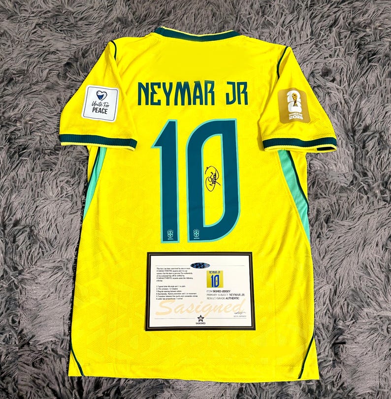 May include: A yellow Brazil soccer jersey featuring the name NEYMAR JR and the number 10 in dark green with a black signature on the number. The jersey includes a Unite For Peace patch, a World Cup 2022 emblem, and a certificate of authenticity.